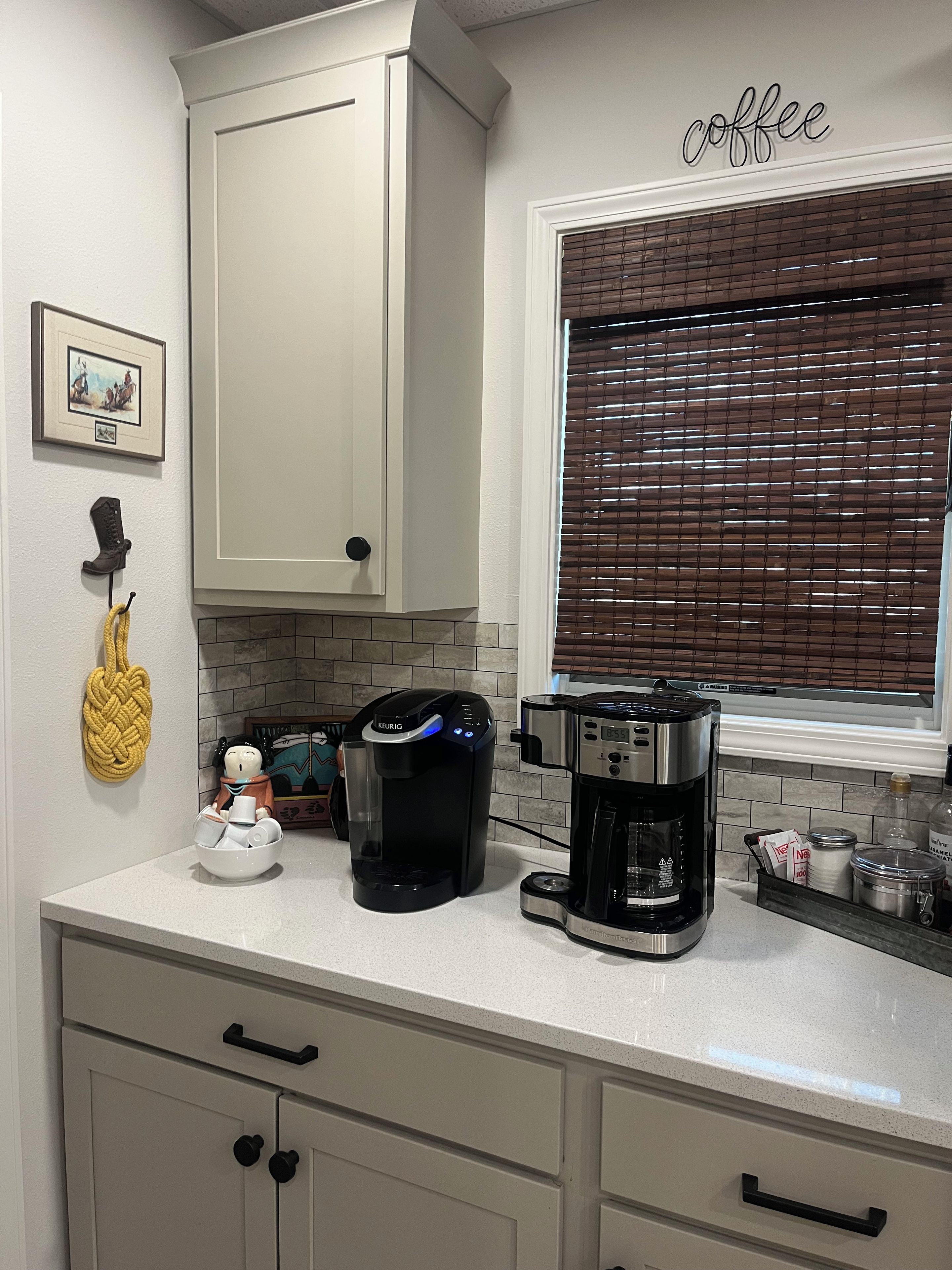 La cuisine est équipée d’une machine à café, d’une machine Keurig et d’une cafetière. Il y a une fenêtre avec des stores, un tableau encadré au mur et une corde jaune qui pend au mur.