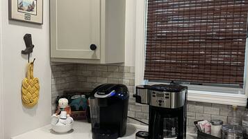 La cuisine est équipée d’une machine à café, d’une machine Keurig et d’une cafetière. Il y a une fenêtre avec des stores, un tableau encadré au mur et une corde jaune qui pend au mur.