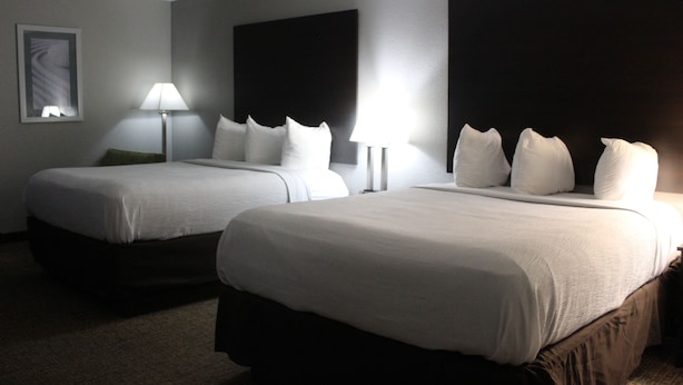 Habitación de hotel con dos camas, cada una con ropa de cama blanca y cabeceros oscuros. Se ve una lámpara de noche individual en una de las camas.
