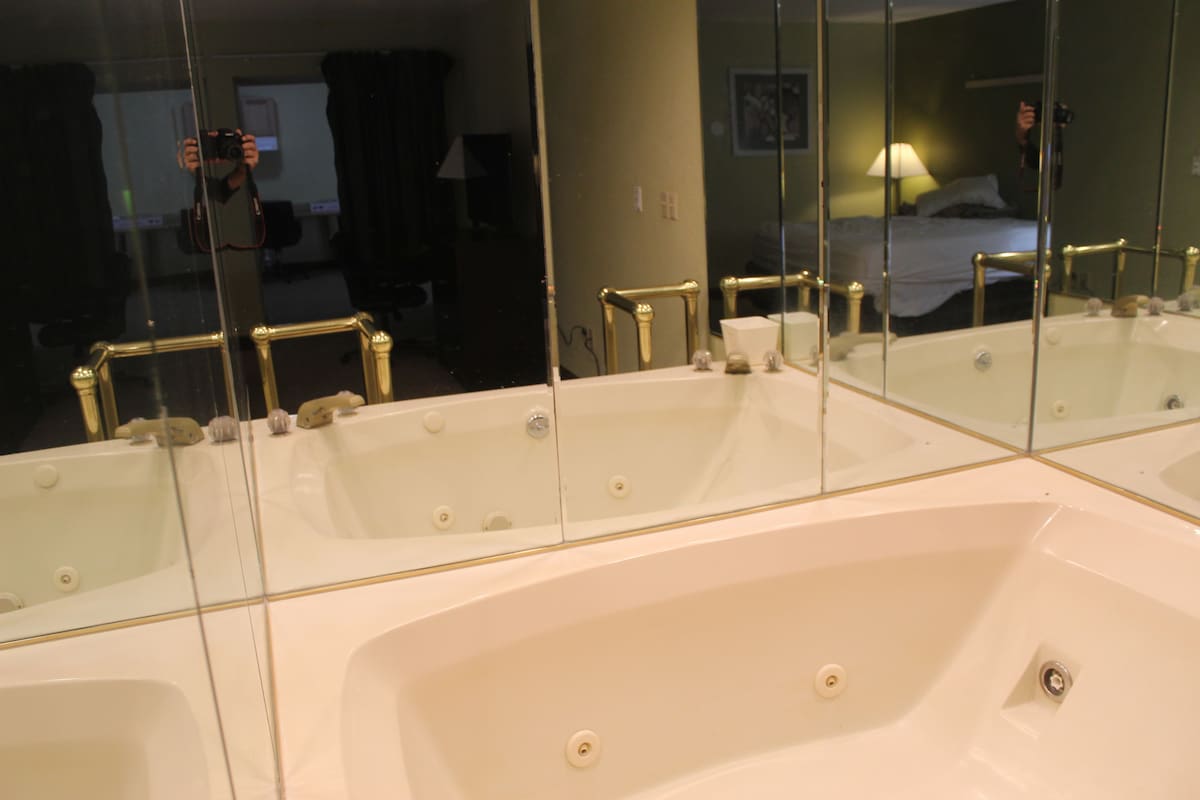 A large white bathtub with gold fixtures, a mirror reflecting a bed and a person taking a photo, and a visible bathroom counter.