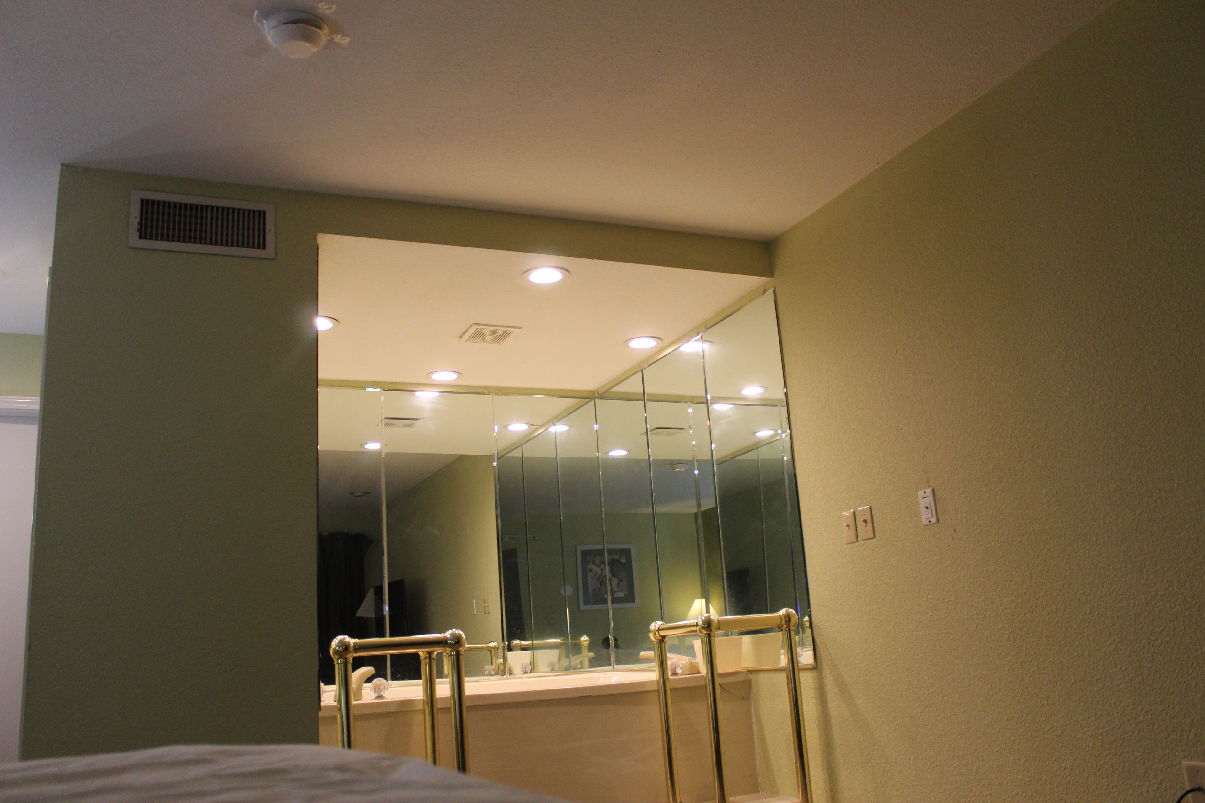 A hotel room with a large mirror, a bed, a window, and a bathroom with a glass partition.