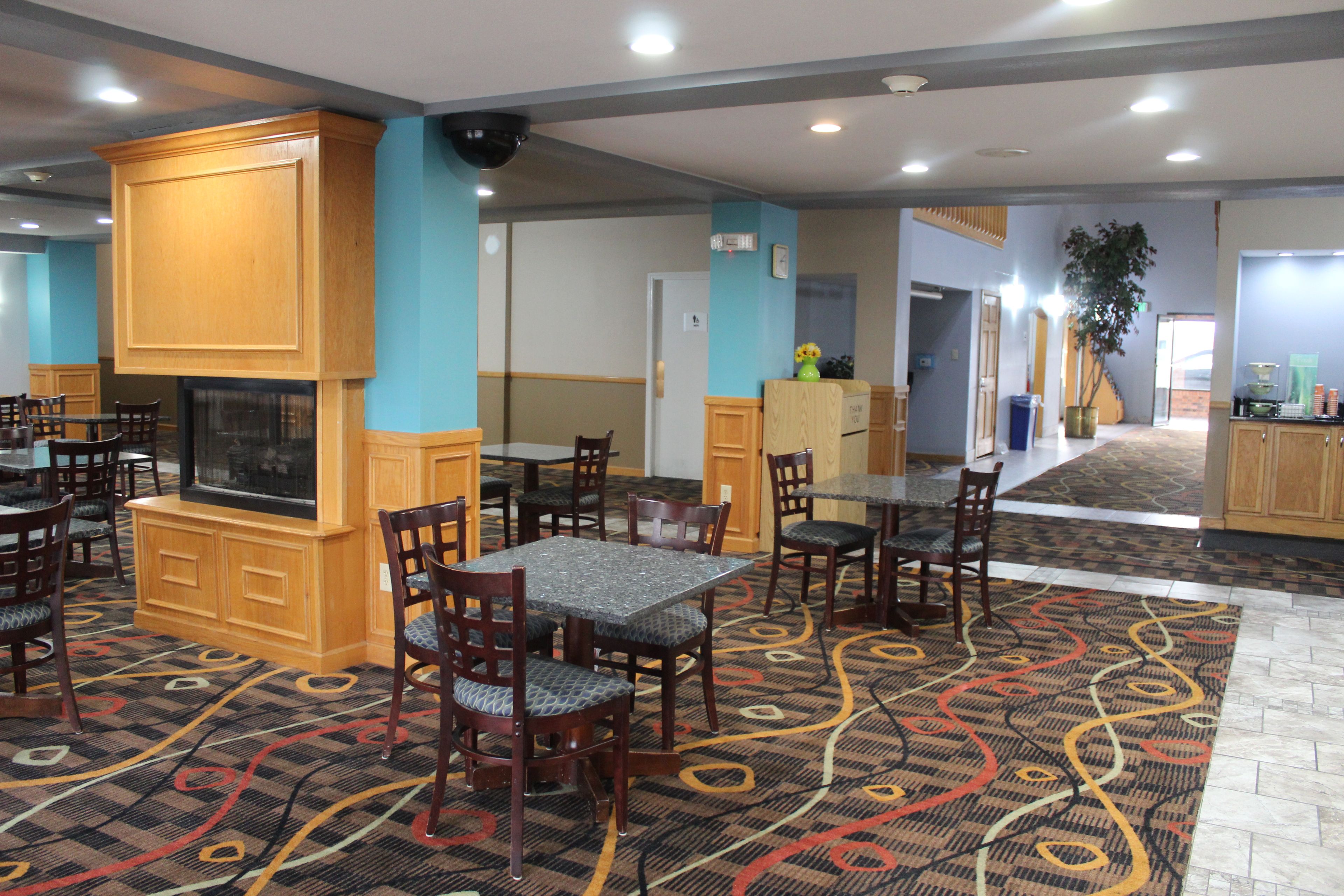 A hotel dining area with multiple tables and chairs, a fireplace, and a patterned carpet.