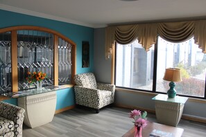 Una habitación con un sillón estampado, un escritorio con tapa de vidrio y un florero, y vistas al exterior a través de una ventana con cortinas.