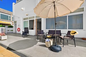 An outdoor seating area with a table and chairs, a large umbrella, and a building with a sign that reads 'MENCEY' in the background. - Azahara Playa Suites (Los Cristianos)
