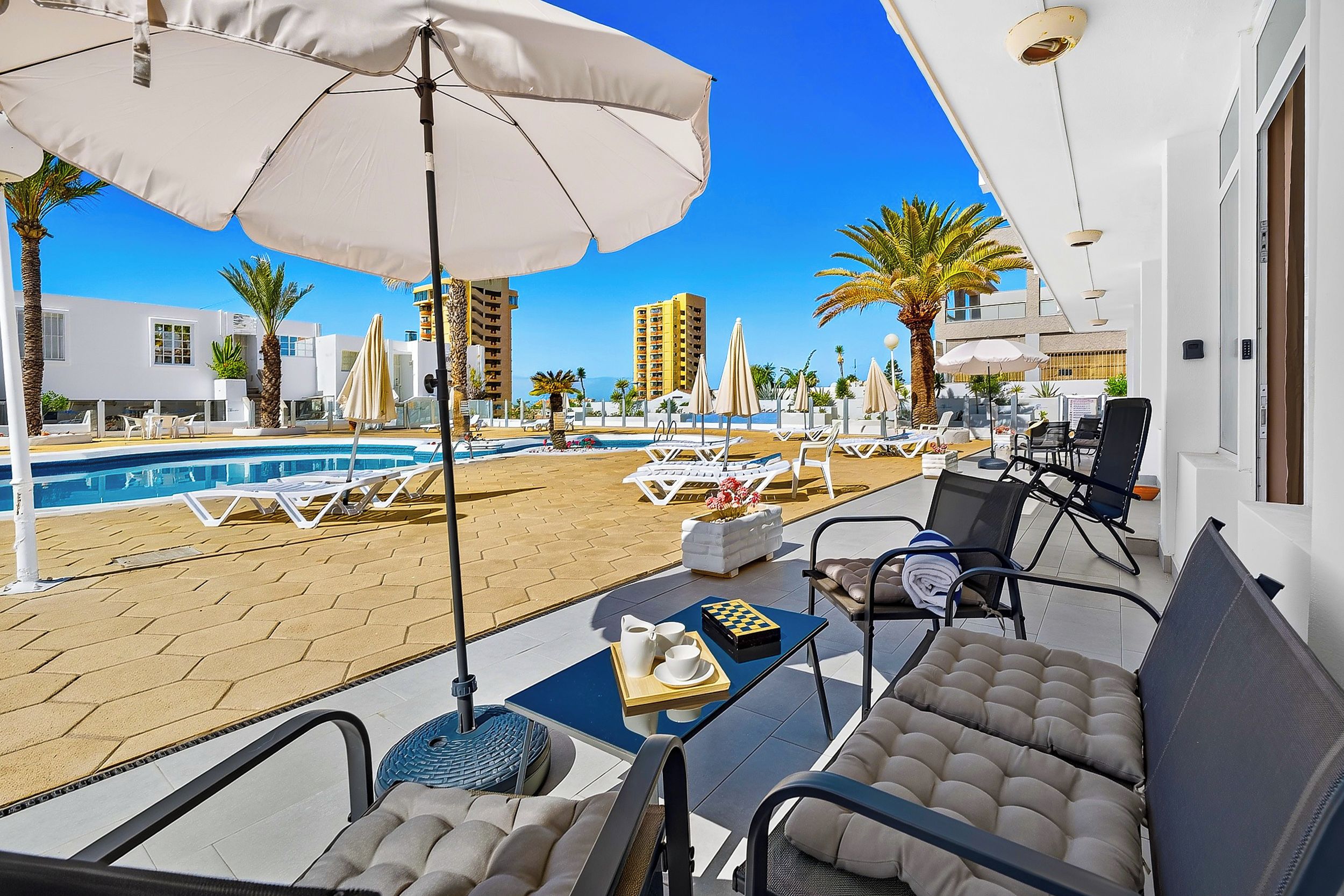 Un espace aménagé au bord de la piscine, avec des chaises longues, des parasols et une vue sur la plage et les bâtiments.