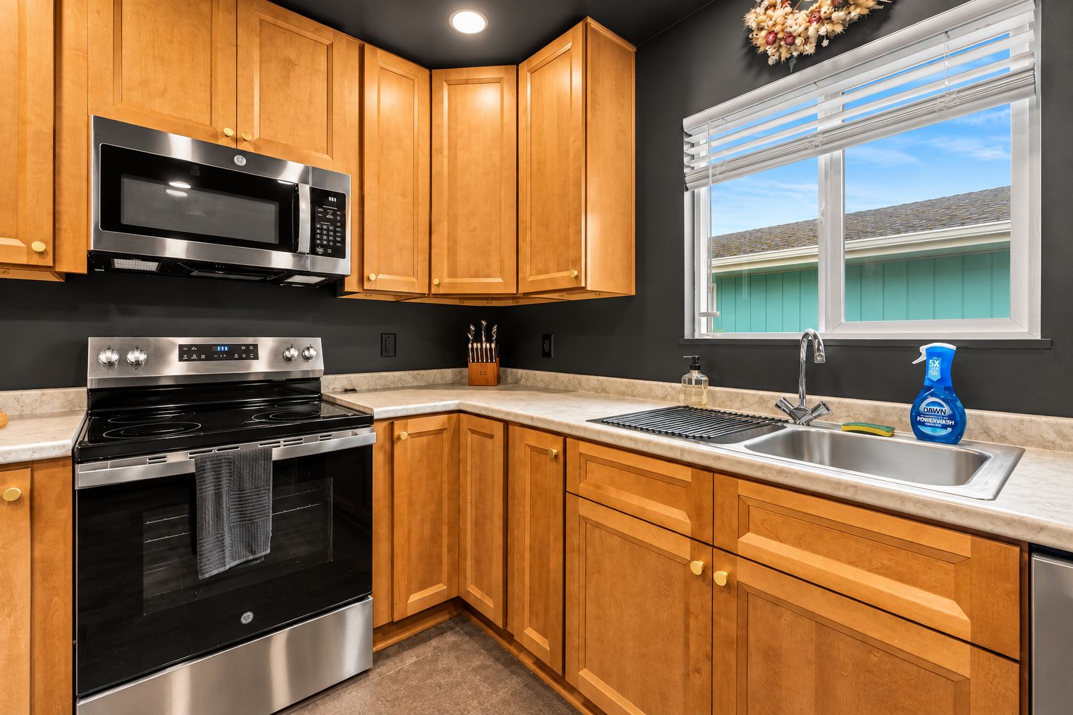 A kitchen with wooden cabinets, a stainless steel microwave, an oven, a sink, and a window with a view of a house.