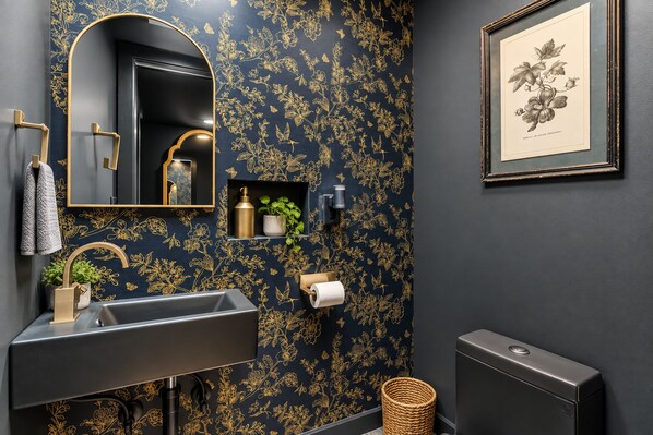 A bathroom with a dark wall, a gold floral pattern, a sink with a gold faucet, a framed botanical illustration, and a small shelf with toiletries. - Octopus Palace (Crescent)