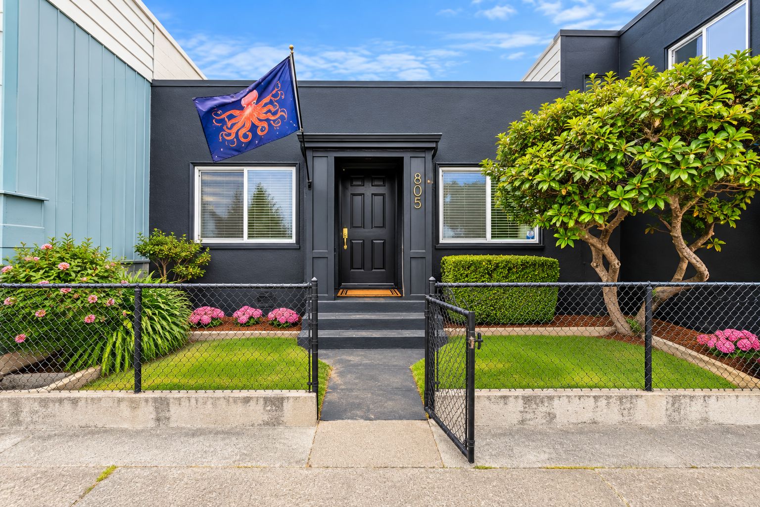A building with a black door, a blue flag with an octopus, and a well-maintained front yard with hedges and flowers.
