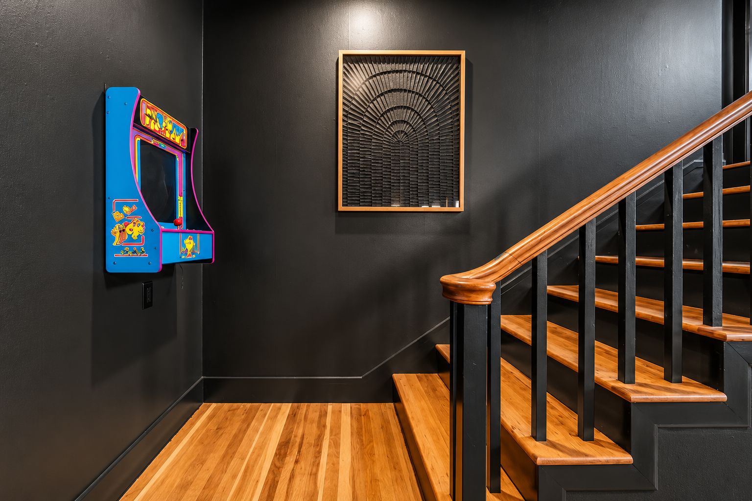 Un escalier avec des marches en bois et des rampes noires, une borne d’arcade fixée au mur et une œuvre d’art abstraite encadrée.