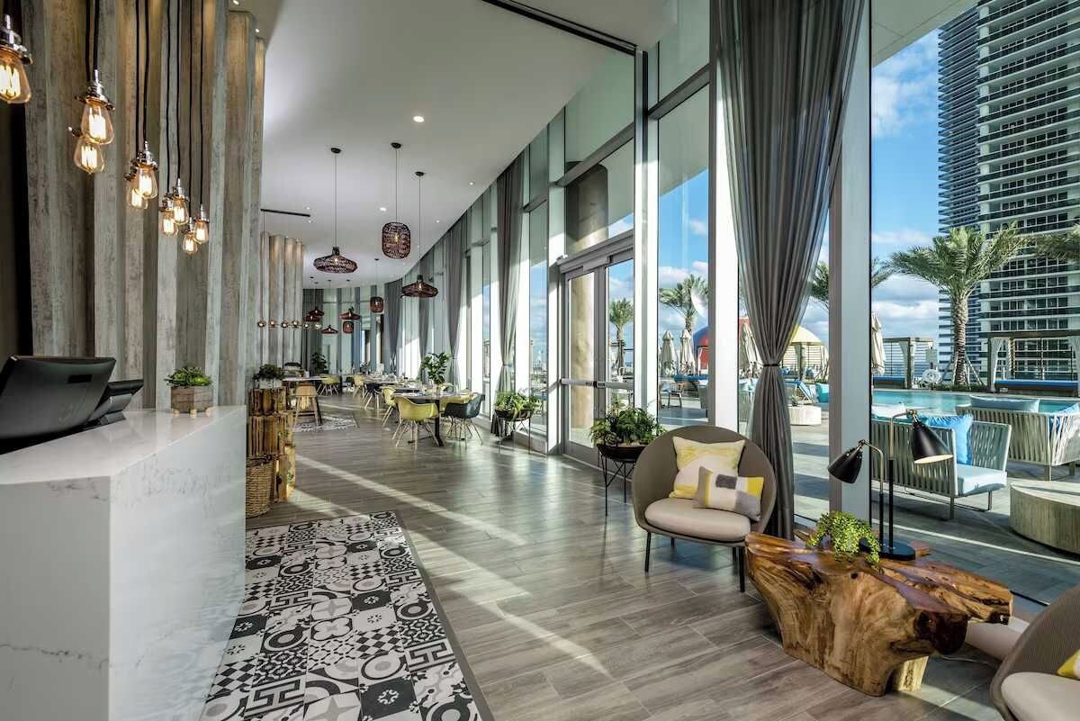 A modern hotel lobby with a long reception desk, seating area, and large windows offering views of a pool and palm trees.