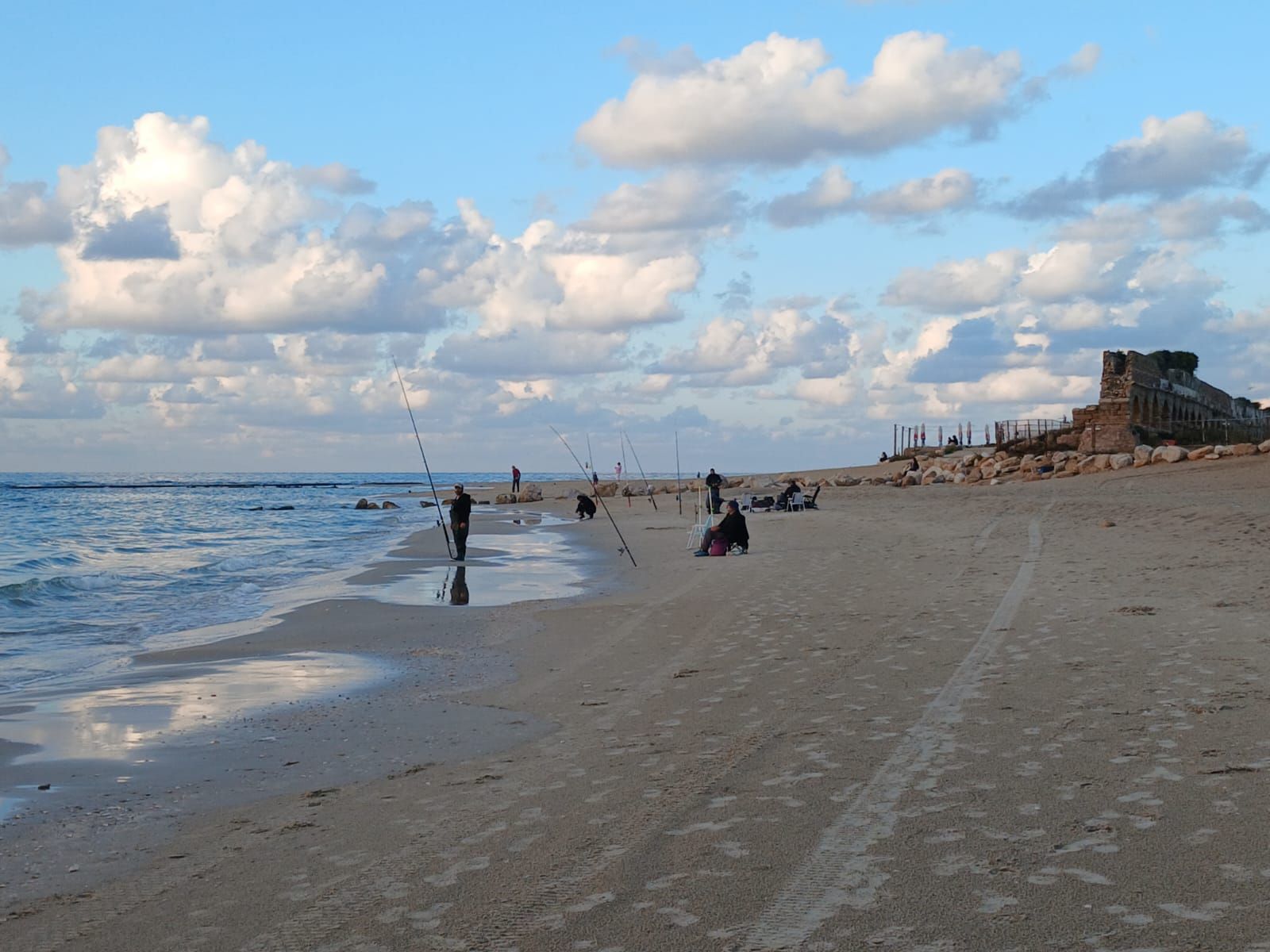 A beach with people fishing, a clear sky, and a historical building in the background.