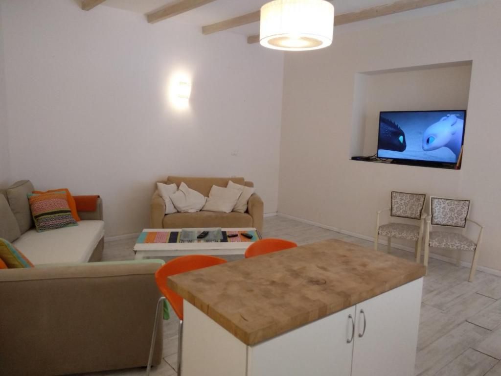 A living room with a sofa, a coffee table, a TV mounted on the wall, and a small kitchen area with a counter and cabinets.