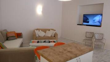 A living room with a sofa, a coffee table, a TV mounted on the wall, and a small kitchen area with a counter and cabinets.
