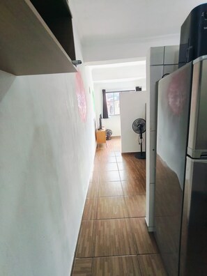 A hallway with a tiled floor, a refrigerator, and a fan in the background.