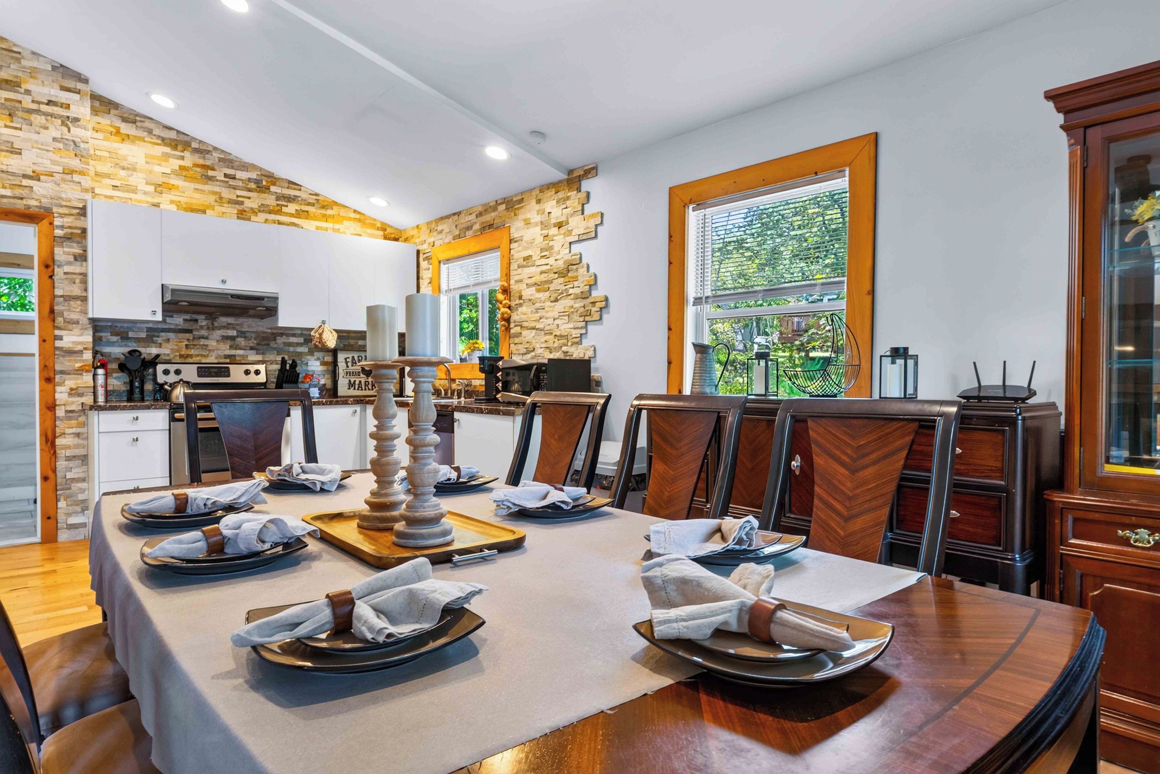 Un comedor con una mesa larga dispuesta para una comida, una cocina al fondo y un ventanal con vista a follaje.