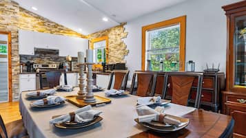 Un comedor con una mesa larga dispuesta para una comida, una cocina al fondo y un ventanal con vista a follaje.