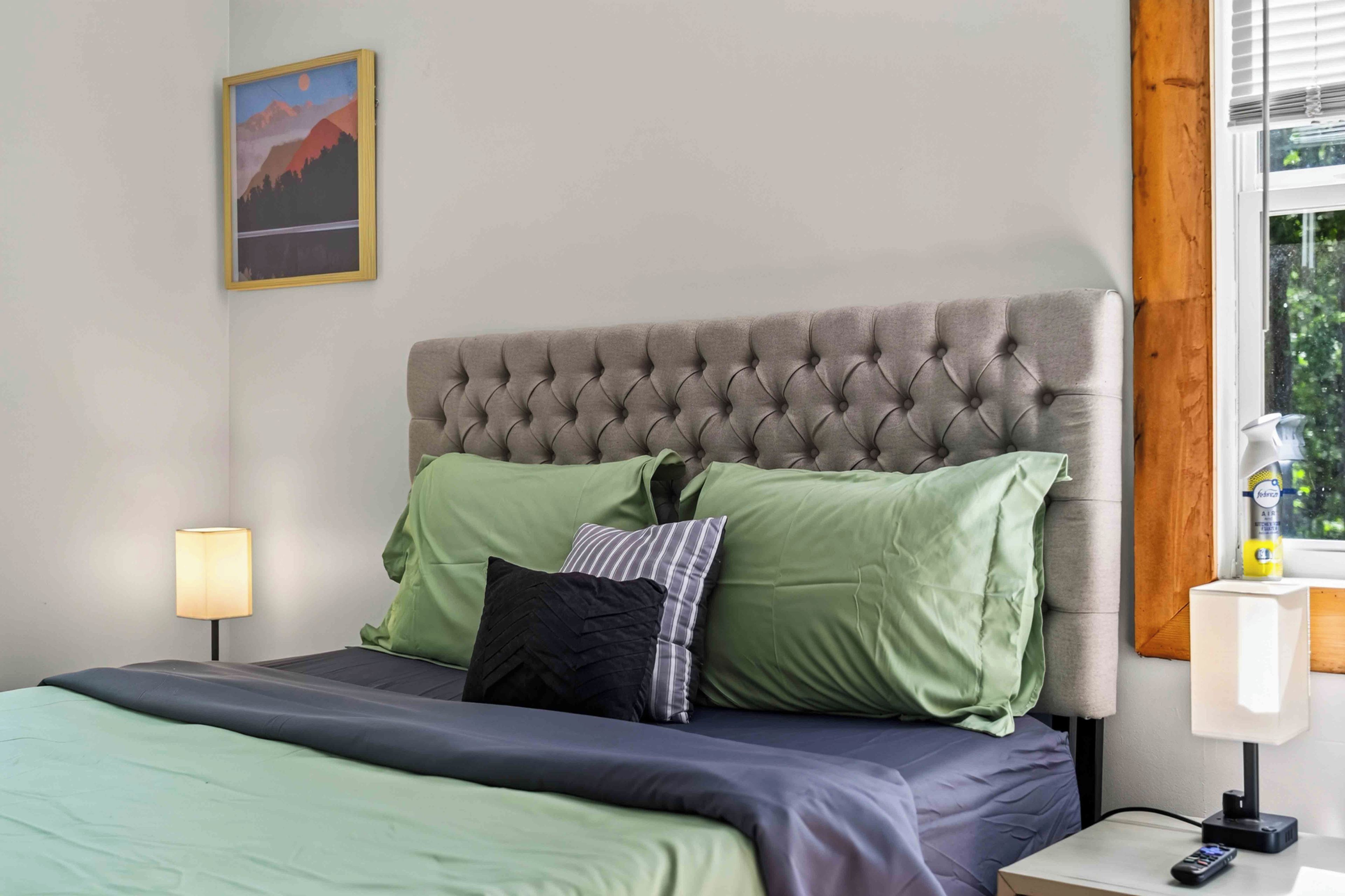 A bedroom with a tufted headboard, green and black pillows, a bedside lamp, and a painting on the wall.