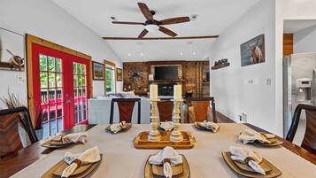A dining area with a long table set for a meal, a ceiling fan, and a kitchen in the background.