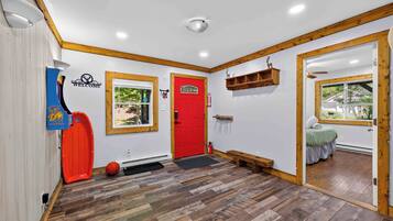 Una habitación con puerta roja, un banco de madera, un estante pequeño y una ventana con vistas al verdor.