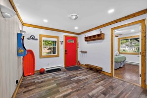 Una habitación con puerta roja, un banco de madera, un estante pequeño y una ventana con vistas al verdor.