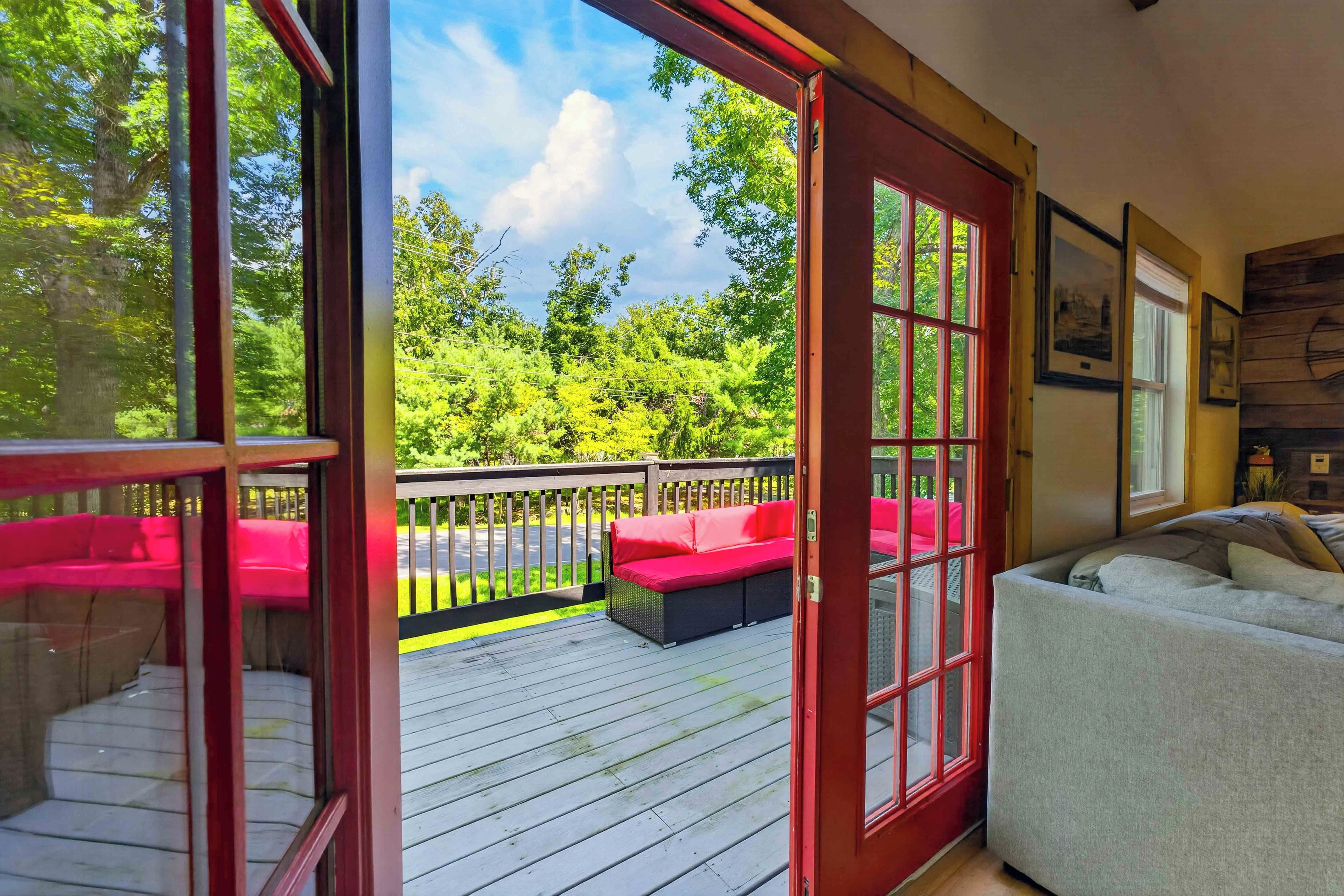 A covered outdoor deck with red-framed glass doors, a red sofa, and a grey sofa inside a room with a view of a green landscape.