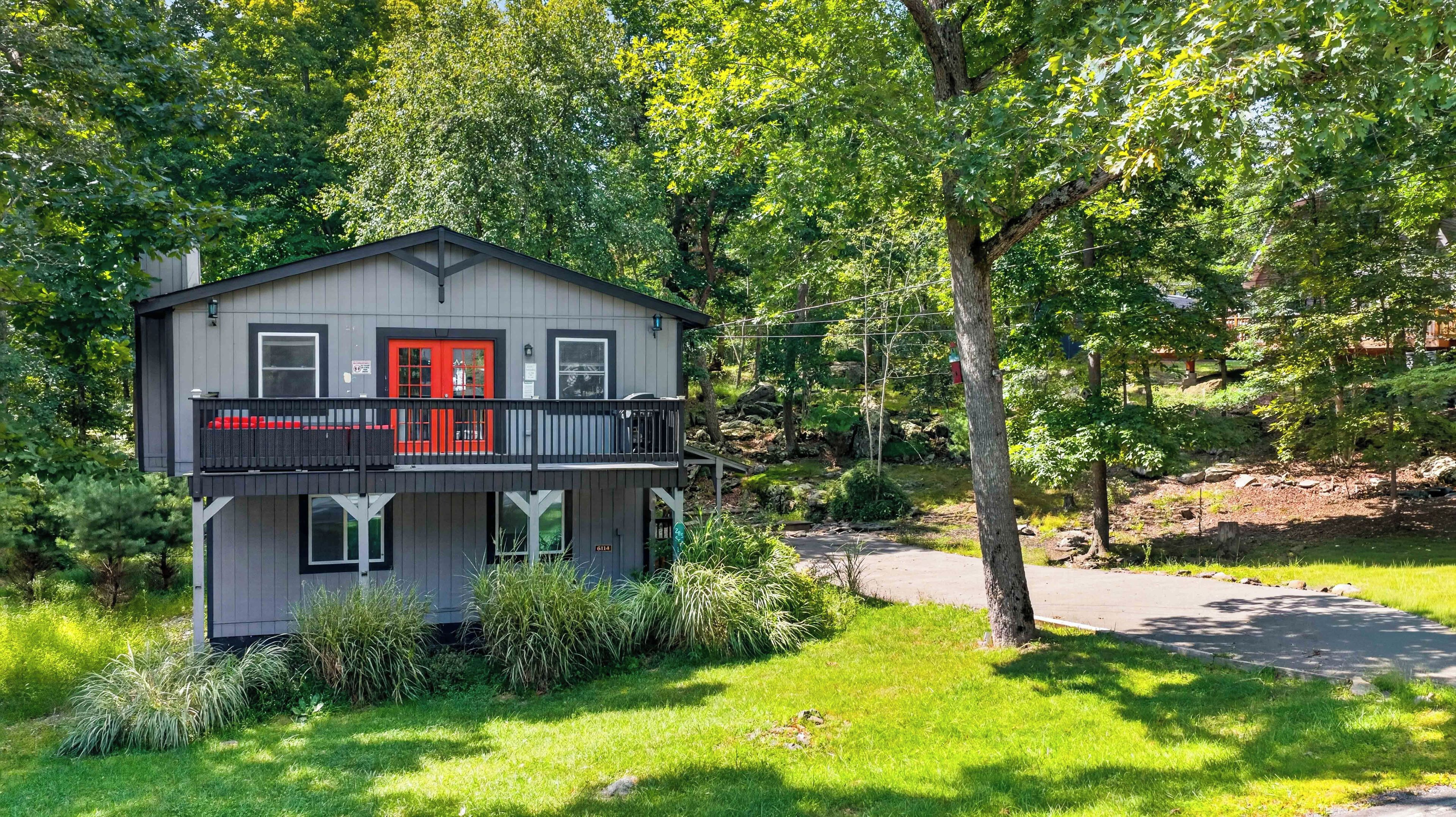 A two-story house with a red door, a balcony with black railings, and a surrounding lush green landscape.
