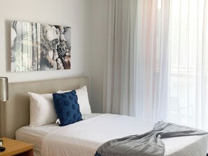 Ein Hotelzimmer mit Bett, Nachttisch, Lampe und einem Fenster mit durchscheinenden Vorhängen.