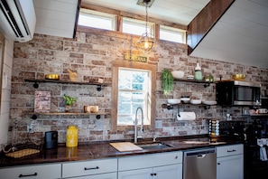 A kitchen with brick walls, wooden shelves, a built-in microwave, and a window with a view. - Cozy Cabin Glacier Getaway - Fireplace & Firepits (Kalispell)