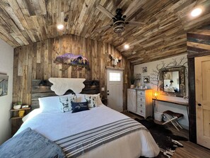 A bedroom with a wooden ceiling, a bed with patterned bedding, a dresser, a mirror, and a ceiling fan. - Cozy Cabin Glacier Getaway - Fireplace & Firepits (Kalispell)