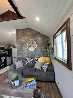 A living room with a gray sofa, a coffee table with books, a decorative wall piece, and a window with blinds. - Cozy Cabin Glacier Getaway - Fireplace & Firepits (Kalispell)
