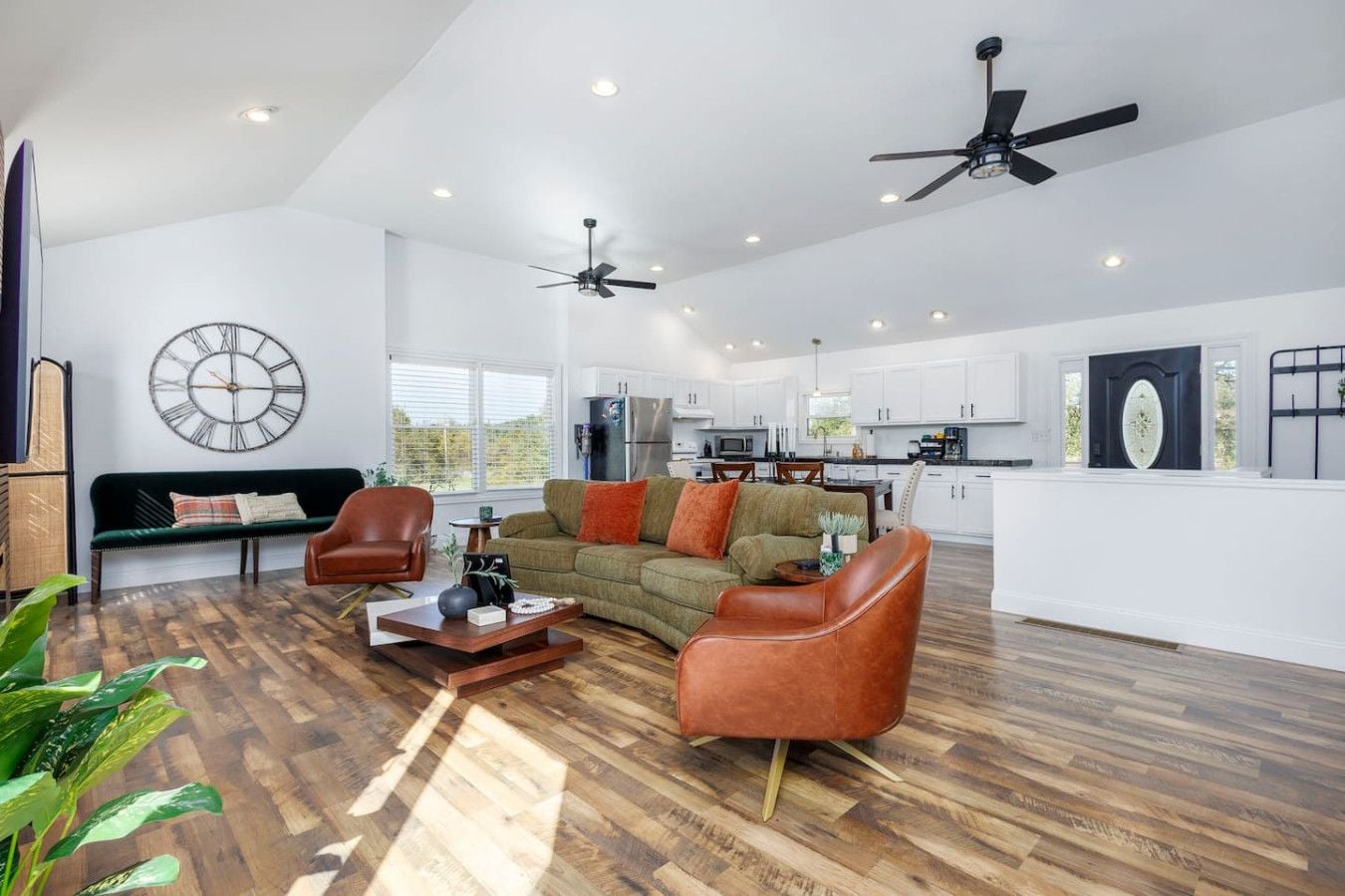 A spacious living area with a sofa, chairs, a coffee table, and a dining table. There are ceiling fans, a kitchen in the background, and a large clock on the wall.