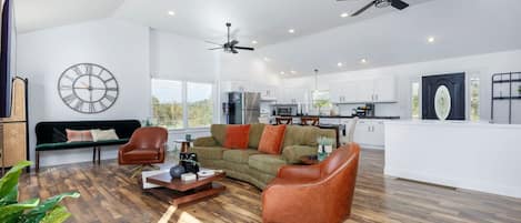 A spacious living area with a sofa, chairs, a coffee table, and a dining table. There are ceiling fans, a kitchen in the background, and a large clock on the wall.