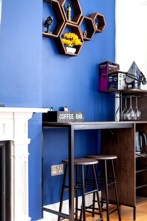 Pared azul con repisa hexagonal que sostiene flores amarillas y figuritas, un letrero de cafetería y una cocina con cafetera y utensilios.