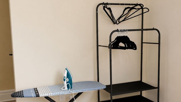 A room with a wooden floor, a metal ironing board with an iron, and a metal shelf unit with a hanging rack.