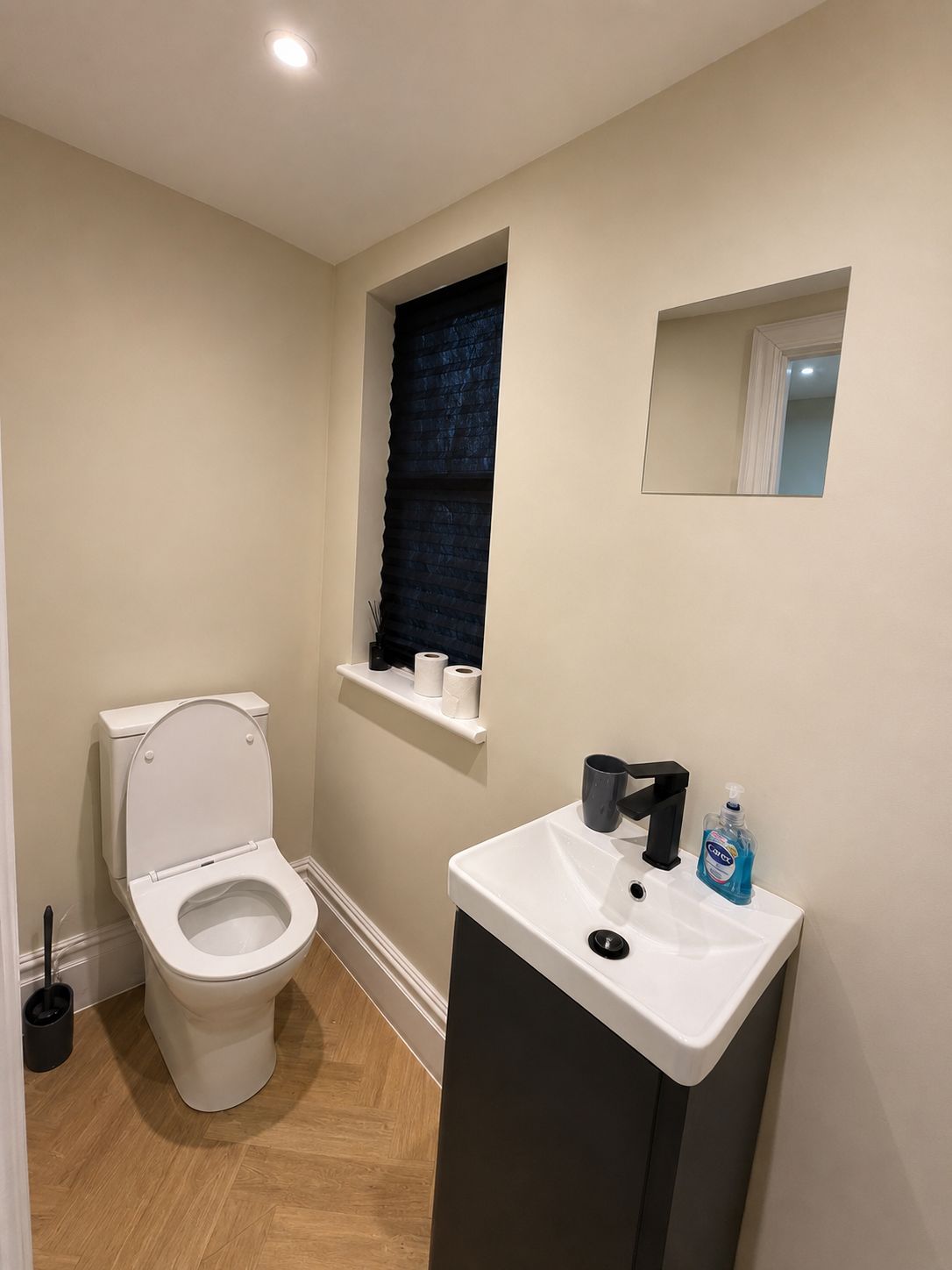Een kleine badkamer met een toilet, een wastafel, een raam met een donker zonnescherm en een spiegel.