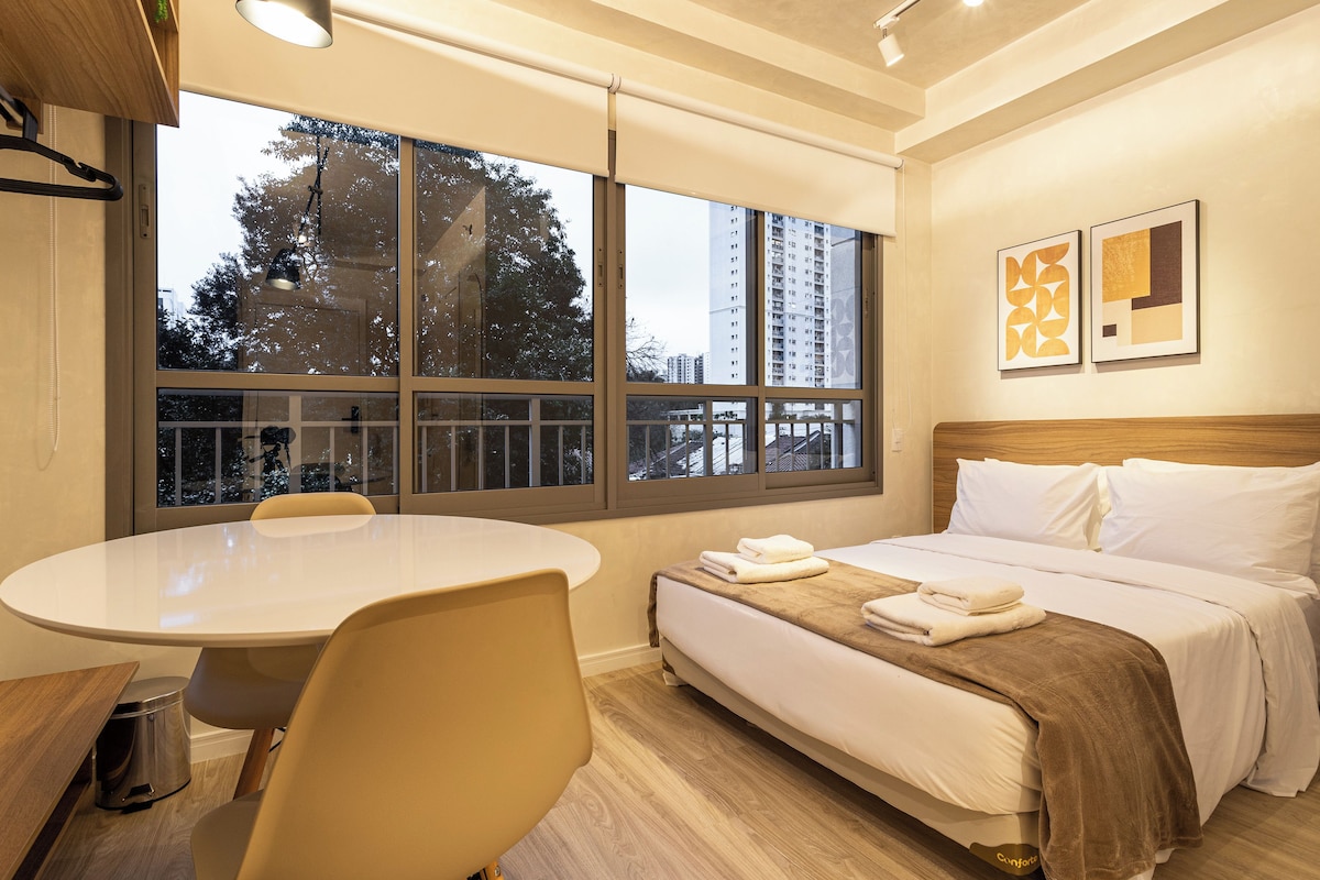 A hotel room with a bed, a round table, a chair, and a window with a view of buildings and trees.
