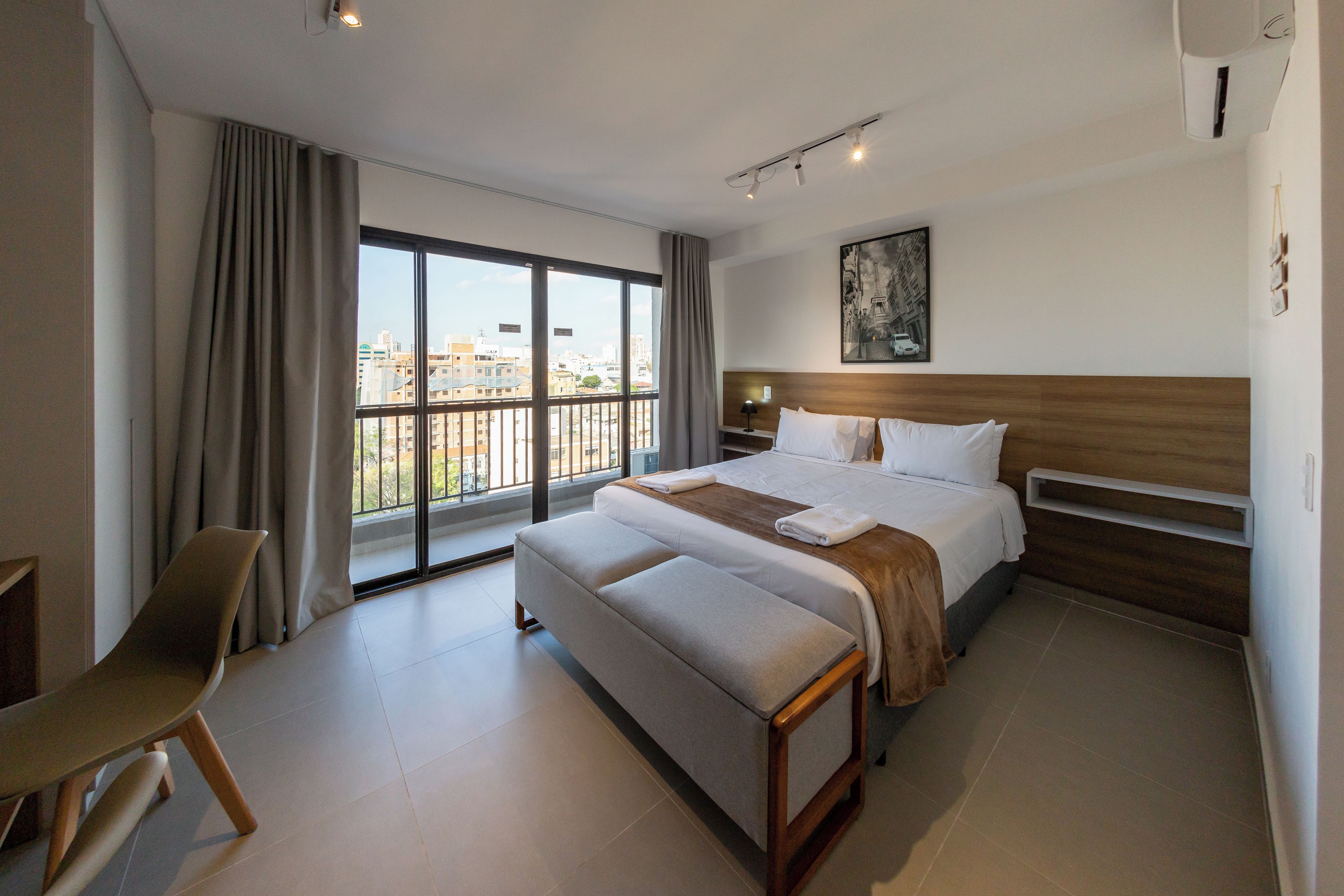A hotel room with a large bed, a smaller sofa, a desk, and a view of the city through large windows.