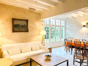 Quarto com sofá branco, mesa de jantar com cadeiras, um quadro na parede e janela com vista para o verde.