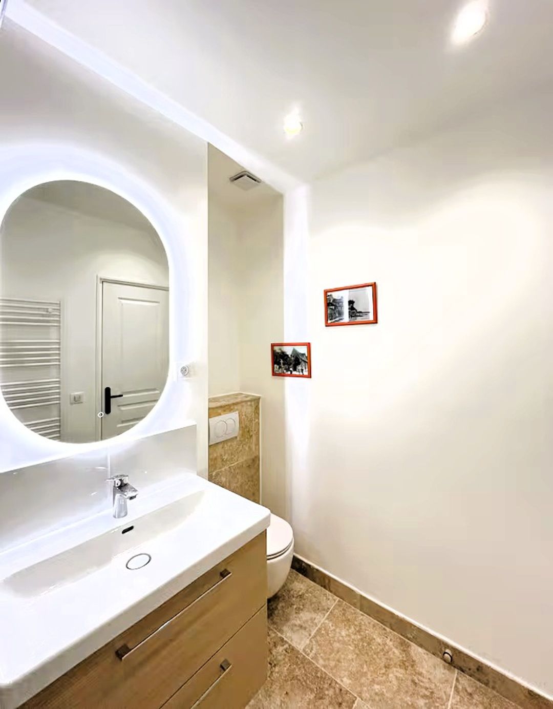 Une salle de bain avec un meuble-lavabo blanc, un miroir rond, des toilettes et deux tableaux encadrés au mur.