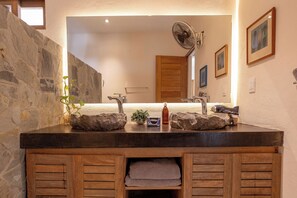 A bathroom with a stone sink, wooden vanity, and a mirror reflecting a fan and a door.