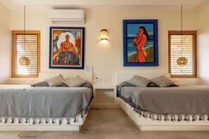 A room with two beds, each with a headboard, a wall-mounted air conditioner, and two framed artworks above the beds.