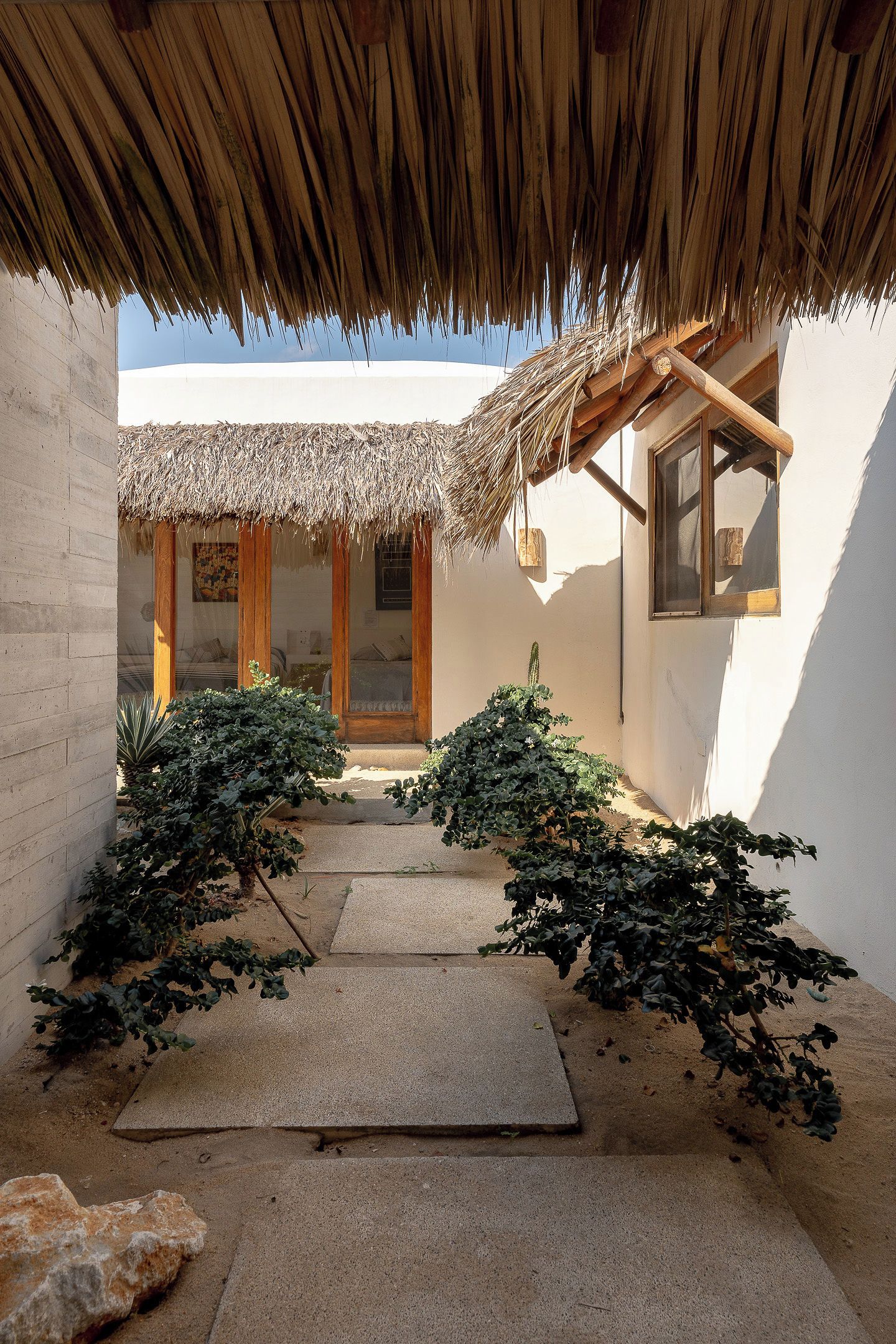 Un patio con una estructura de techo de paja, un sendero de concreto y un edificio con puerta y ventanas de madera.