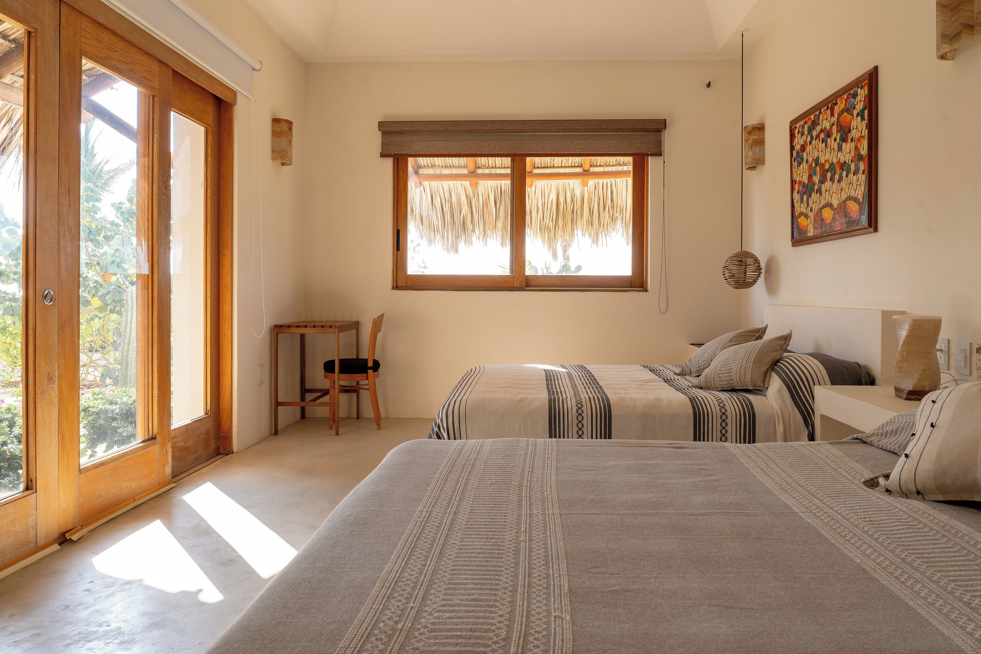 Un dormitorio con dos camas, una silla, un ventanal con tejado de paja y un cuadro en la pared.