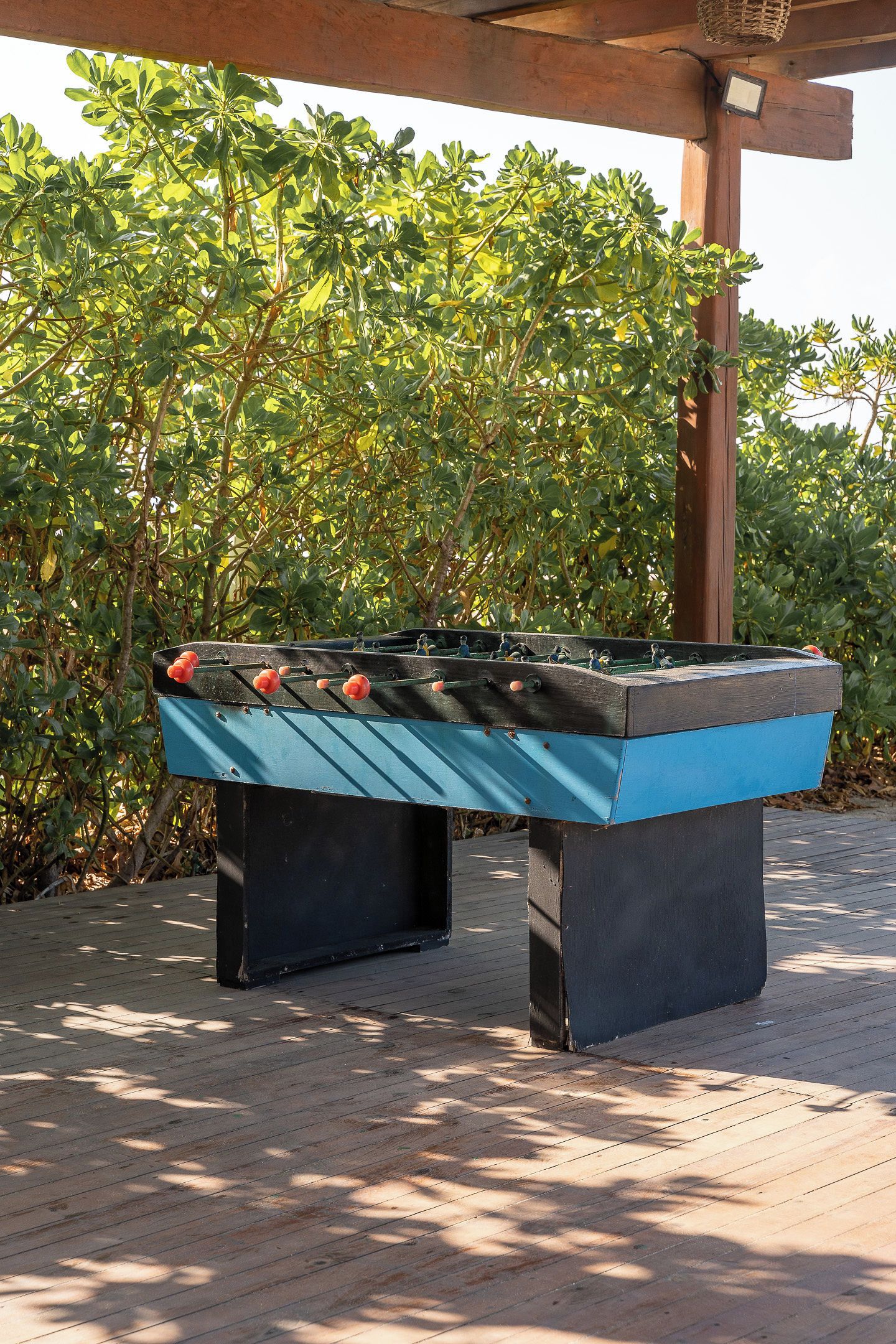 A foosball table outdoors with a wooden structure overhead and dense foliage in the background.