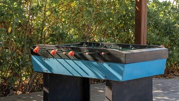A foosball table outdoors with a wooden structure overhead and dense foliage in the background.