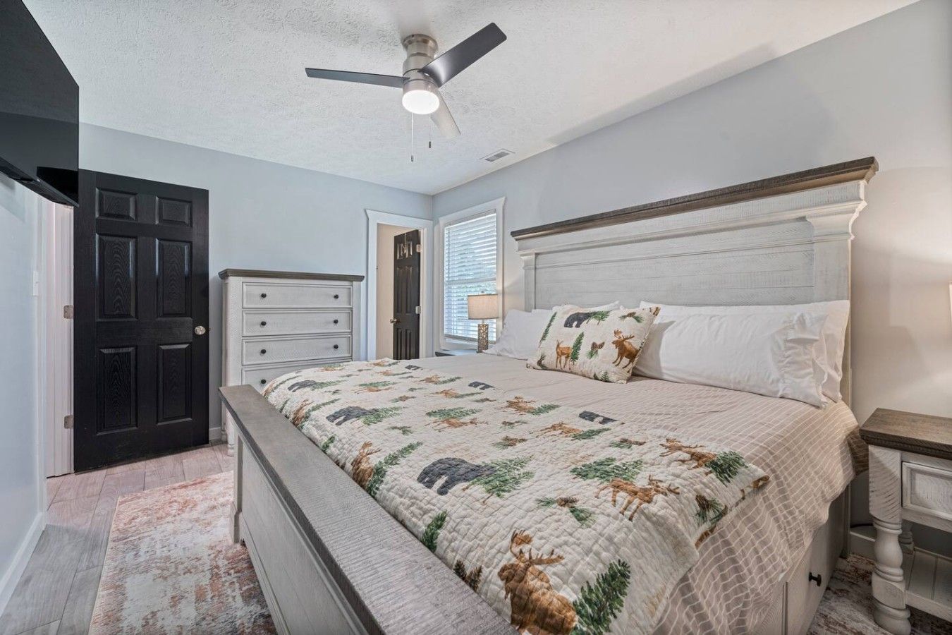 Une chambre à coucher comprenant un lit, une table de chevet, une commode, un ventilateur de plafond et une porte.