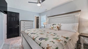 Une chambre à coucher comprenant un lit, une table de chevet, une commode, un ventilateur de plafond et une porte.