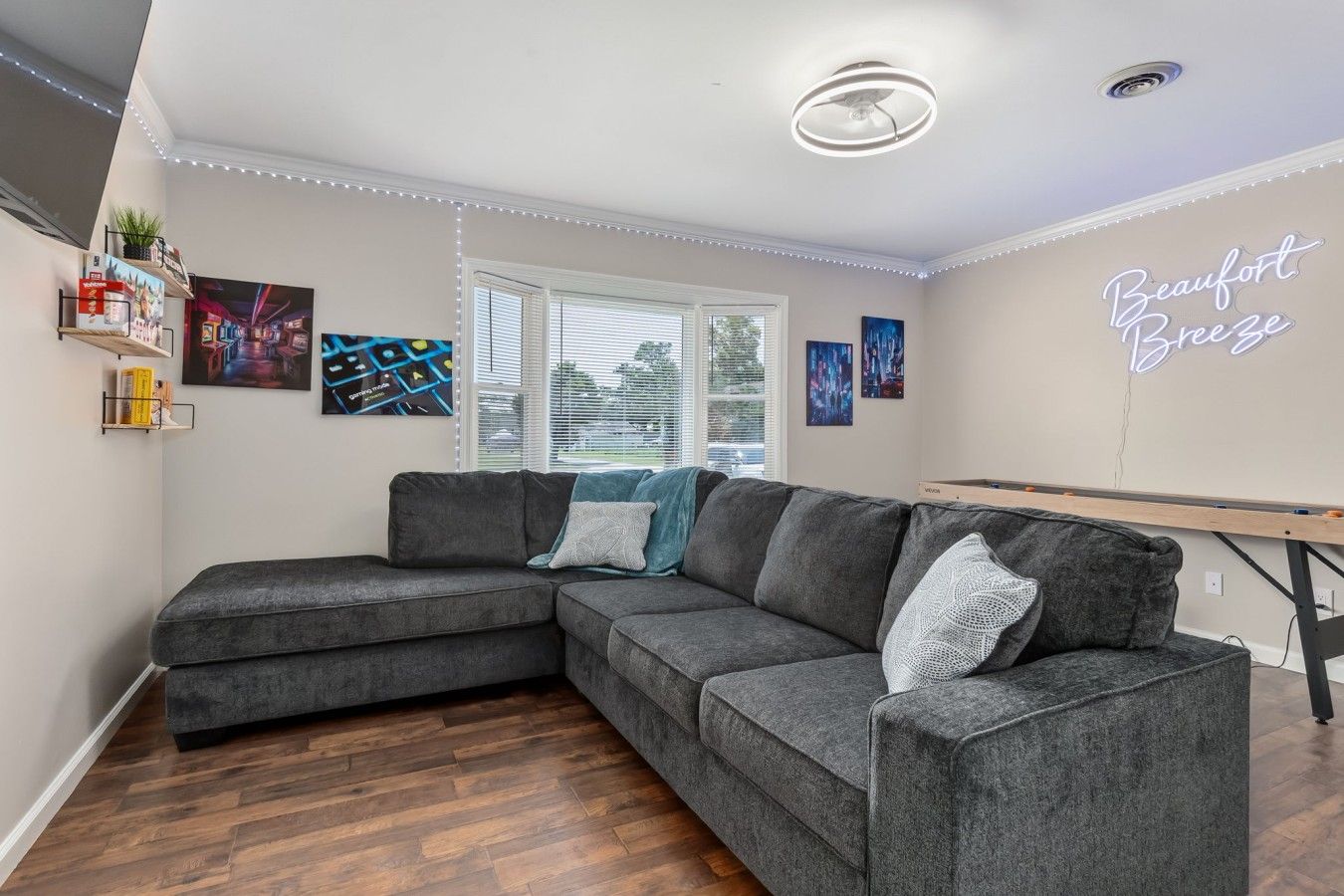 A modern living room with a grey sectional sofa, wooden flooring, and a neon sign on the wall.