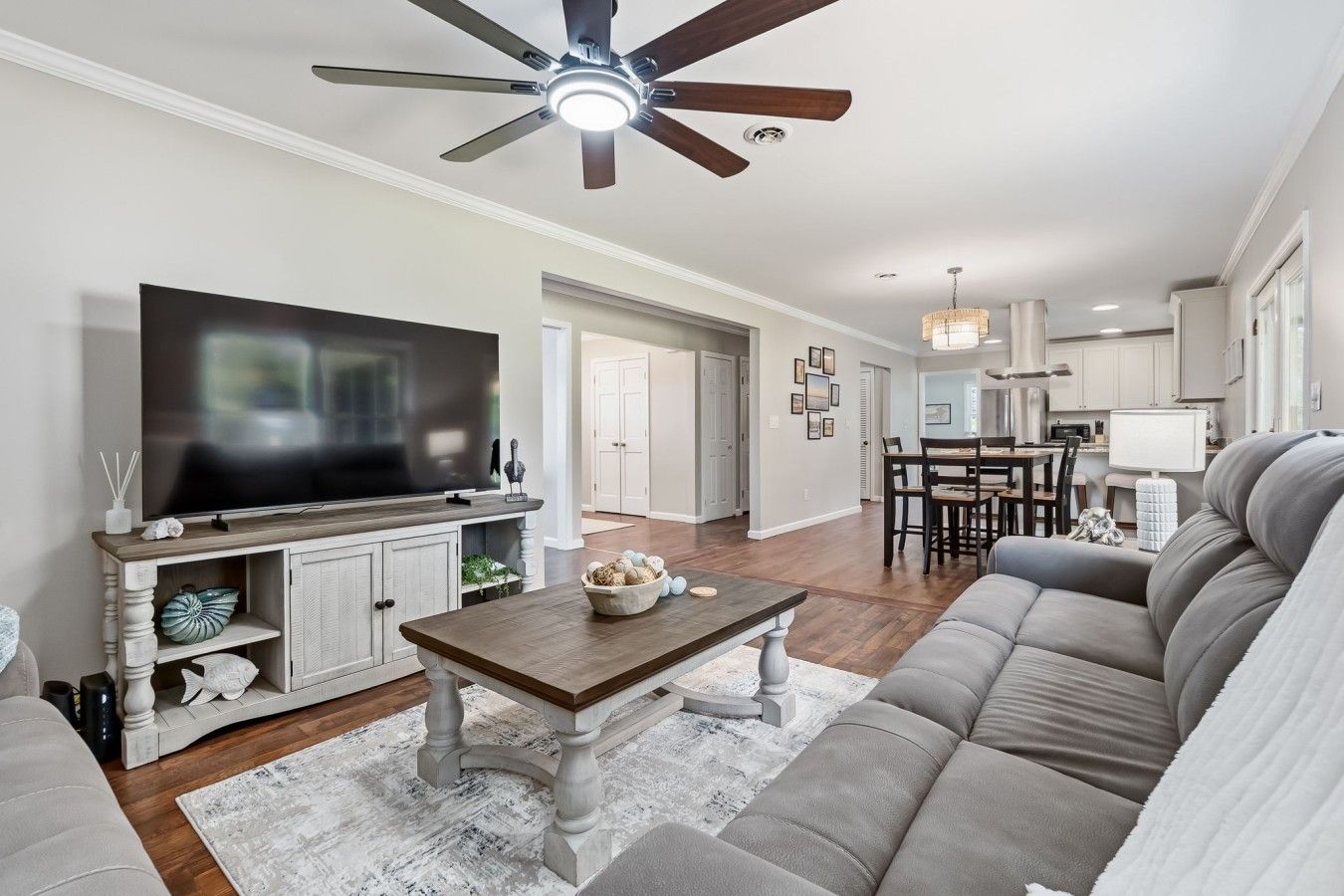 A modern living room with a flat-screen TV, a ceiling fan, a dining area, and a kitchen in the background.