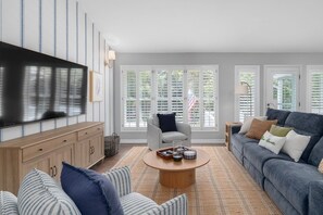 Ein Wohnzimmer mit Fernseher, einer Couch, einem Sessel, einem Couchtisch und großen Fenstern mit Rollläden.